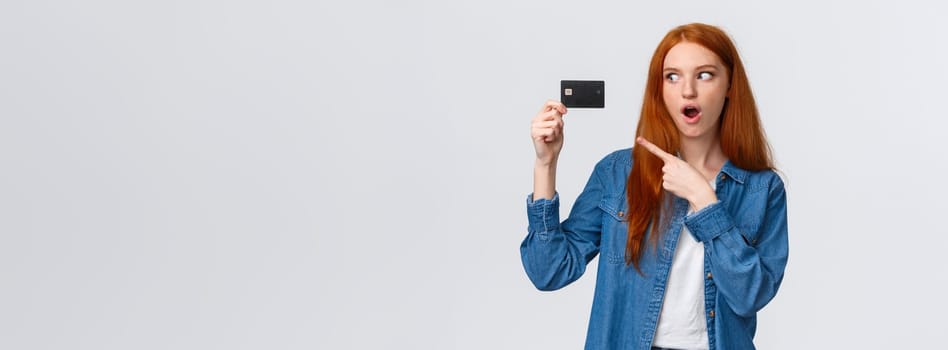 Discounts, shopping and banking concept. Waist-up impressed, thrilled cute redhead girl in denim shirt holding credit card, pointing at it with fascinated impressed eyes, standing white background.