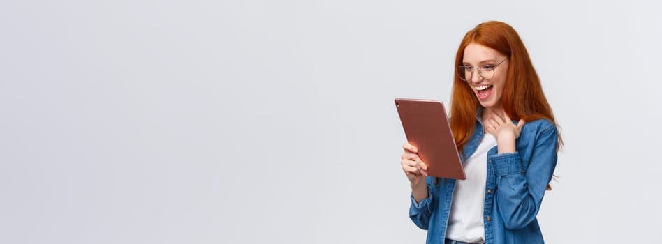 Excited, happy and surprised cheerful redhead woman messaging with friend using gadget, holding digital tablet, touch chest pleased and amazed, laughing out loud looking device screen.