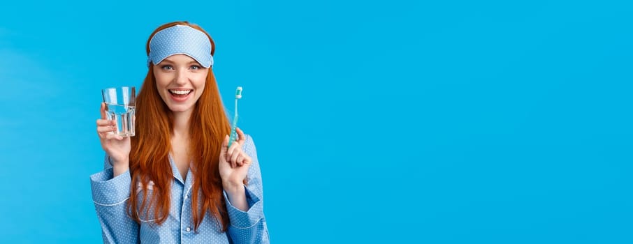 Advertising, hygiene and people concept. Feminine cute redhead girl feeling upbeat and enthusiastic start day right, drinking water, holding glass and toothbrush, wear nightwear and sleep mask.