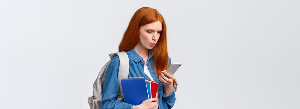 What is that. Confused and displesaed. intense troubled redhead teenage, college girl with backpack and notebooks walking university and reading strange text on smartphone, white background.