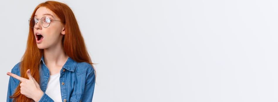 Cool take a look. Close-up portrait fascinated and excited cute redhead woman in glasses checking out awesome banner, pointing and looking left with opened mouth, say wow, white background.