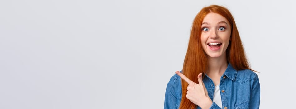 Close-up portrait excited happy ginger girl, foxy hair and blue eyes, smiling upbeat with amazement and joy, pointing finger left, attend amusing cool party, telling about it, white background.