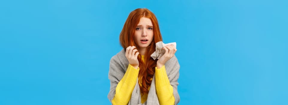 Dizzy, uneasy sad redhead female wearing scarf to get warm, catching cold, sneezing and having runny nose, using napkin, looking camera with tired exhausted expression, feel fatigue, blue background.