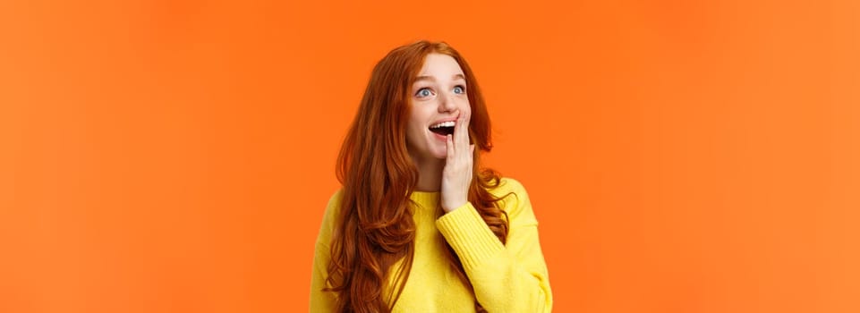 Fascinated and amiring cute redhead woman looking impressed and excited at upper left corner, smiling see breathtaking view, cover opened mouth palm thrilled, losing speech happy, orange background.