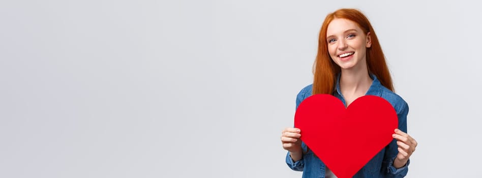 Valentines day, romance and teenagers concept. Lovely and cute redhead girlfriend asking wanna go prom together with big red heart, confessing love, smiling joyfully, white background.