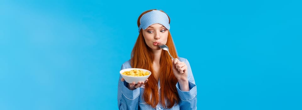 So tasty. Cute and tender feminine girl starting day with healthy diet, eating delicious cereals, licking spoon and staring plate tempting and eager eat it, standing blue background.