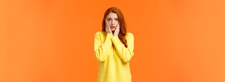 Concerned and anxious redhead girl forgot take chicken out freezer before mom came home, grab face, gasping scared and troubled, dont know what do, standing tensed and afraid, orange background.