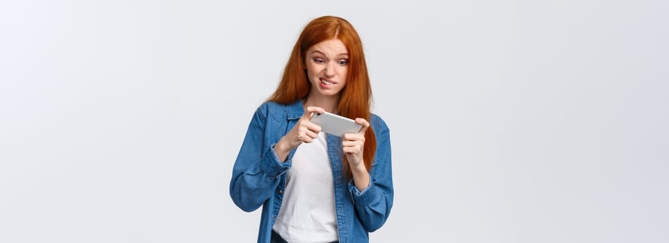 Girl having intense fighting round in game, tap screen and trying beat friends score in app. Excited good-looking redhead female student wasting time playing smartphone arcade, white background.