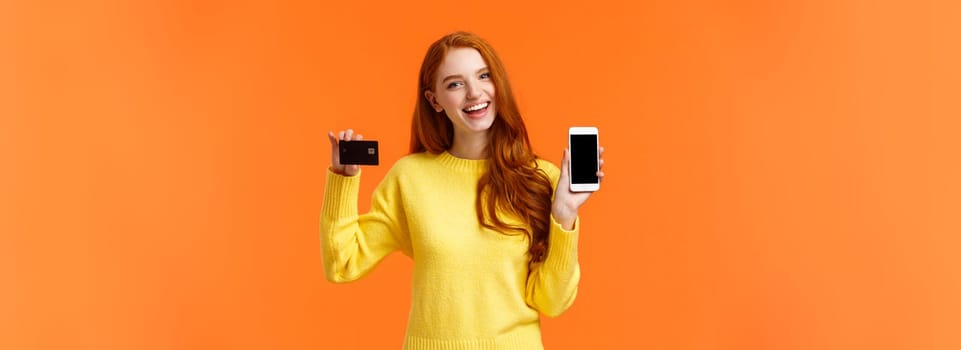 Shopping, online and finance concept. Cheerful young redhead female opened bank account, holding smartphone and credit card, smiling, recommend use online shopping application.