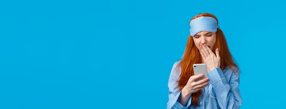 Gadgets addication, lifestyle and beauty concept. Lovely redhead girl going sleep and scrolling feed smartphone, yawning squinting feeling tired, wearing pyjama and sleep mask, blue background.