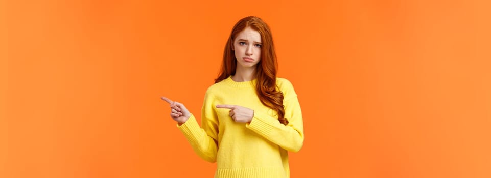 Sulking moody and sad cute redhead girlfriend pointing right and frowning, standing unhappy with bad mood, envy someone, begging boyfriend book tickets for winter ski-resort, orange background.