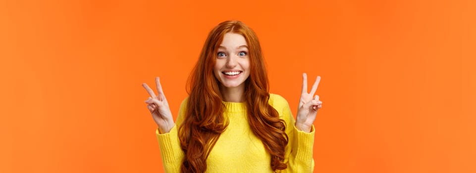 Waist-up portrait lovely and cheerful pretty redhead woman showing peace gestures, smiling camera, travel abroad during winter holidays posing near sightseeing, orange background.