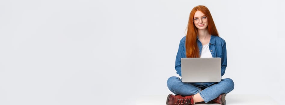 Education, modern lifestyle and college concept. Attractive carefree redhead female student sitting with legs crossed on floor, using laptop, working at coworking space, white background.