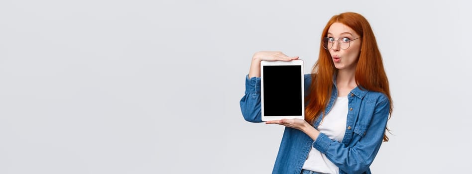 Amused, excited good-looking redhead woman in denim shirt, folding lips in wow sound, looking amazed camera, holding digital tablet, showing device screen, standing white background.