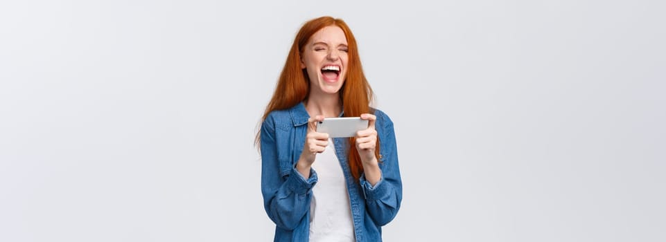 Carefree good-looking millennial redhead girl having fun, watching favorite vlogger, laughing over hilarious video online, holding smartphone horizontally, standing white background joyful.