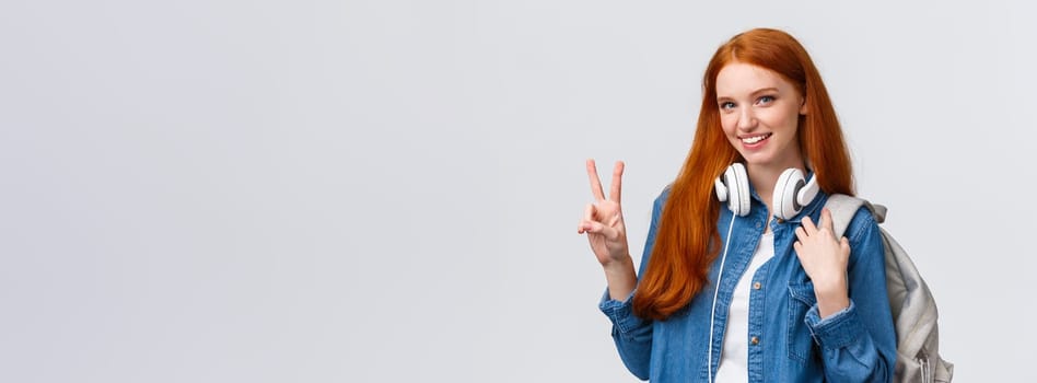 Lovely charismatic caucasian redhead girl with backpack, headphones over neck, showing peace sign and smiling delighted and joyful camera, standing white background, heading part-time job.