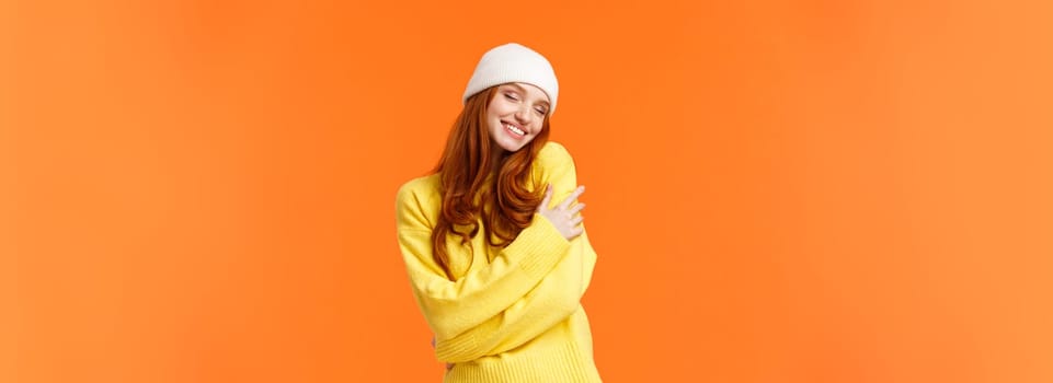 Tenderness, winter holidays concept. Charming romantic redhead woman in soft sweater, white beanie, close eyes and embrace own body, hugging herself, smiling cheerful, orange background.