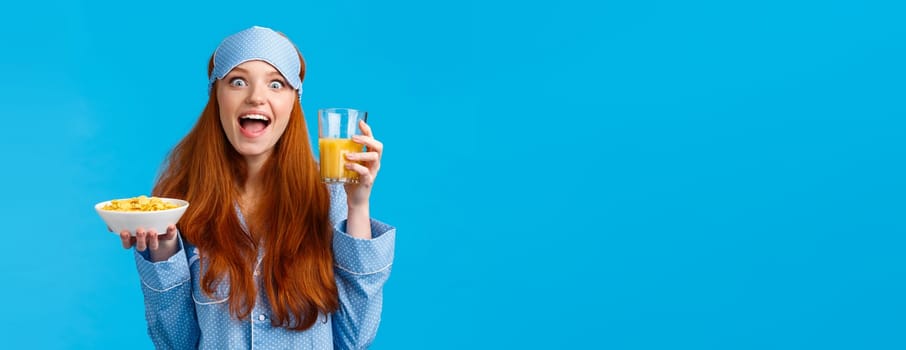 Excited european cute girl with red ginger hair, foxy haircolor, smiling amused and funny gazing camera, holding fresh orange juice and cereals, eating morning breakfast, standing blue background.