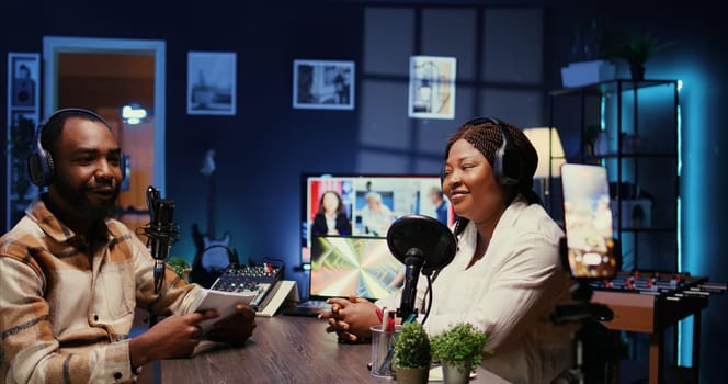 African american host using smartphone on tripod to record entertaining discussion with woman during livestream in apartment studio. Presenter uses mobile phone camera to produce comedy podcast