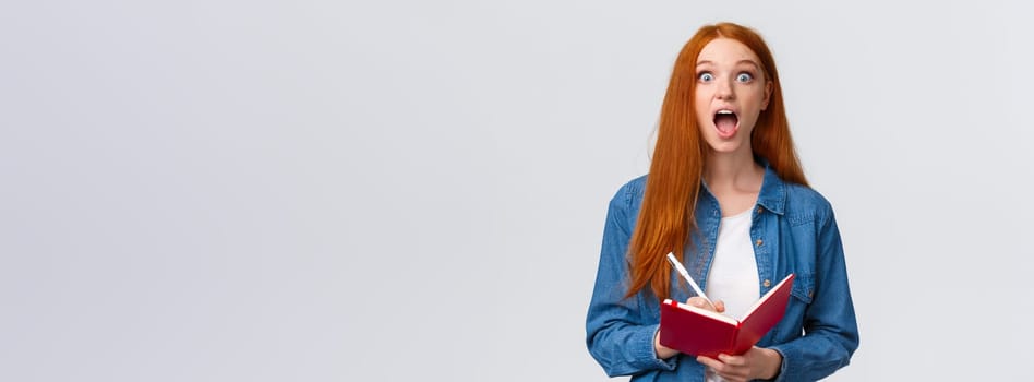 Amused and excited, astonished redhead girl fascinated with amazing lecture giving speech, writing down useful notes, holding notebook and staring thrilled camera, white background.