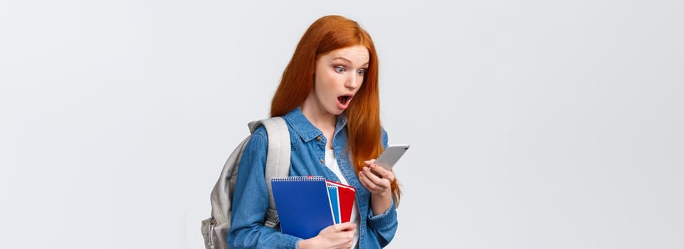 Awesome no classes today. Amazed and excited redhead teenage girl gasping as reading cool message on smartphone, holding backpack notebooks and mobile phone, white background.