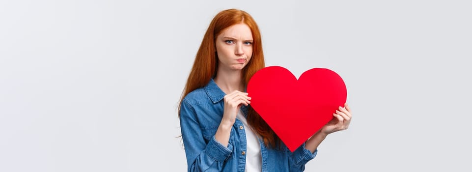 Serious-looking troubled, unsure cute redhead girl thinking what gift buy in addition to valentines day big red heart card, plan romantic date, pouting and frowning camera perplexed.