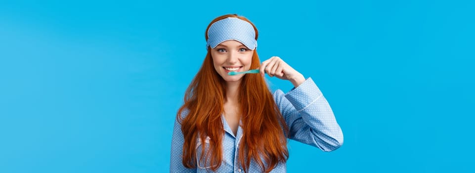 Habits, hygiene and lifestyle concept. Cute feminine redhead european woman in nightwear, sleep mask smiling and brushing teeth with toothbrush, standing blue background.