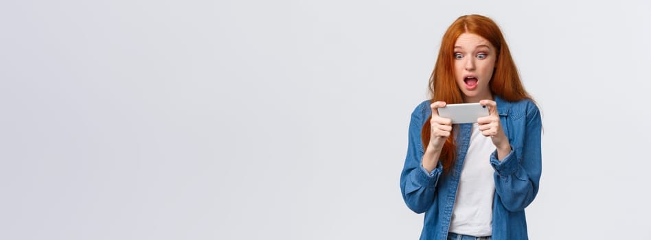 Astonished startled cute redhead woman just won challenge in awesome game, holding smartphone horizonally, playing racing, stare display amazed, cant belive she lucky, white background.