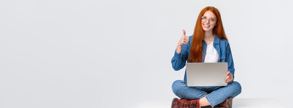 Education, internet and people concept. Female redhead digital nomad, freelance girl in glasses, finished project before deadline, sitting on floor with crossed legs and laptop, show thumb up.