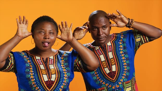 Silly ethnic couple fooling around on camera with comic face, having fun and sticking tongue out in studio. Cheerful people doing funky gesture and enjoying time on set, orange background.