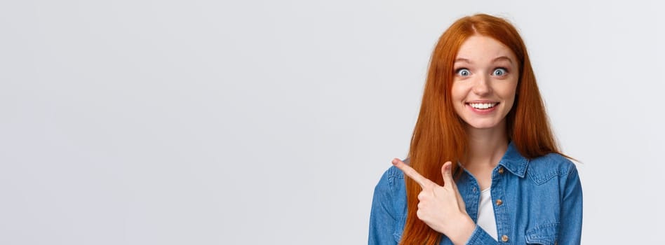 Excited and impressed, curious thrilled smiling redhead girl with long foxy, ginger hair and blue eyes, stare amused camera with wondered grin, pointing finger left, found out awesome news.