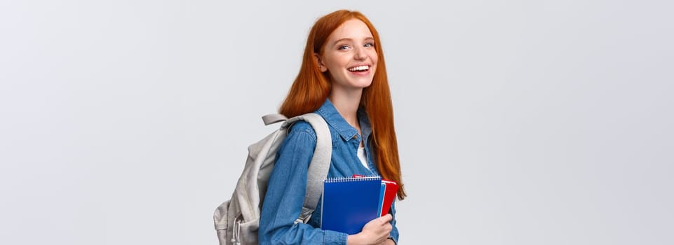 Students, education and studying concept. Lovely charismatic redhead foxy girl with backpack, notebooks and study books, standing white background, smiling joyful looking camera.