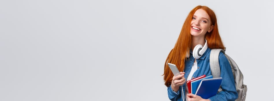 Cheerful, friendly and charismatic redhead woman laughing joyfully, texting friends, chatting with smartphone, enjoying college lifestyle, heading campus with backpack and notebooks.