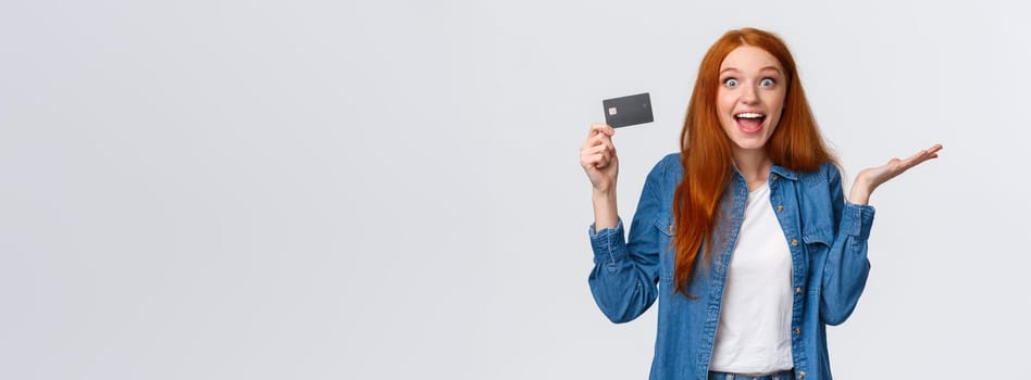 Waist-up portrait surpirsed and cheerful redhead girl talking about bank service, holding credit card smiling amused and excited, raise hand in wonder and thrill, standing white background.