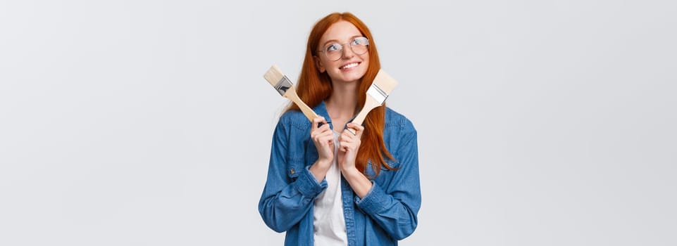 Creativity, imagination and people concept. Good-looking redhead dreamy girl designer, planning to renovate, paint walls in appartment, looking thoughtful and holding paintbrush, white background.