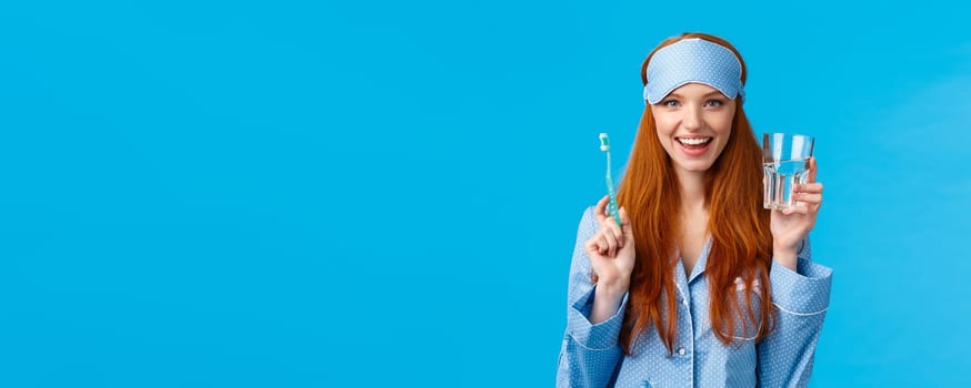 Advertising, hygiene and people concept. Feminine cute redhead girl feeling upbeat and enthusiastic start day right, drinking water, holding glass and toothbrush, wear nightwear and sleep mask.