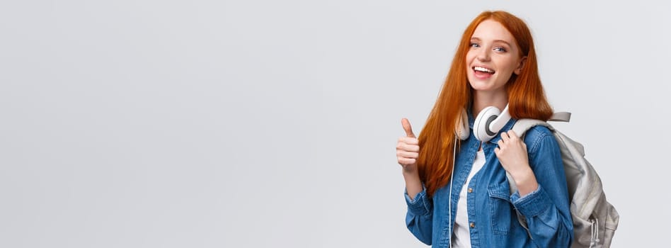 Waist-up portrait joyful cute redhead girl inviting freshmen apply univeristy, got scholarship, smiling showing thumbs-up in approval, recommendation, holding backpack and headphones.