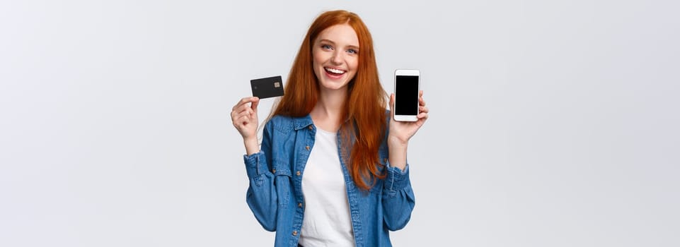 Shopping, application and mobile concept. Waist-up portrait friendly charismatic female blogger, redhead girl promote bank service, showing smartphone or app on display and credit card, smiling.