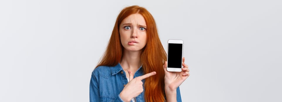 Worried, insecure and alarmed cute redhead girl asking for advice having problems messaging with stranger on dating app, dont know what answer standing nervous pointing finger at smartphone display.