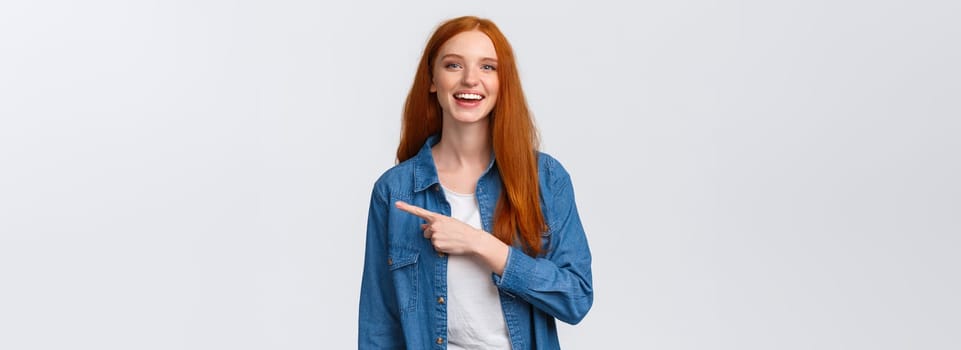 Carefree, friendly and happy outgoing redhead woman in denim shirt, pointing finger right and laughing at camera with beaming smile, standing white background, having discussion, communicate team.