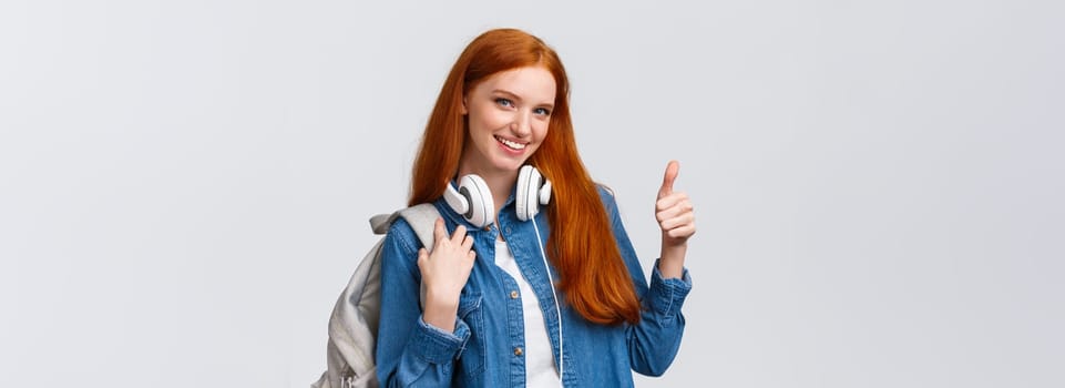 Girl found excellent part-time job after university classes. Attractive redhead woman with backpack, headphones over neck, showing thumb-up and smiling in approval, recommending.