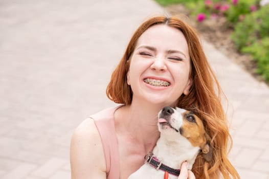 Dog jack russell terrier licks the owner in the face outdoors. Girl with braces on her teeth