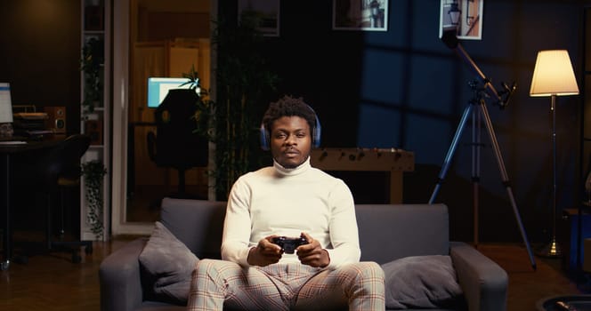 BIPOC man using headphones mic to discuss strategies with teammates while playing videogame in cozy apartment. Player chatting with online strangers while enjoying game on gaming console