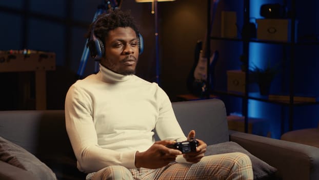 African american man playing videogames on console late at night, struggling to stay awake. Gamer using joypad controller to try and finish singleplayer game level before going to sleep.