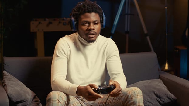 African american man using headphones mic to talk with teammates while playing videogame in cozy apartment. Gamer chatting with online friends while enjoying game on gaming console