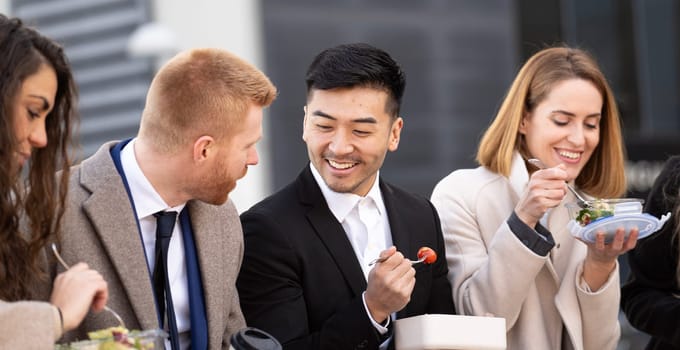 Multiethnic business people eating from lunch boxes outdoors.