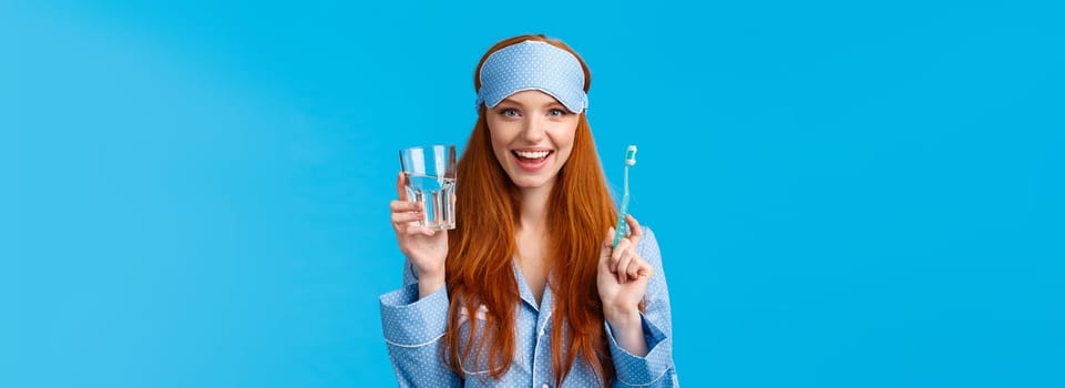 Advertising, hygiene and people concept. Feminine cute redhead girl feeling upbeat and enthusiastic start day right, drinking water, holding glass and toothbrush, wear nightwear and sleep mask.
