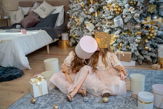 Two cute little girls in beige dresses are sitting under the Christmas tree, putting boxes on their heads. Children under the Christmas tree with gift boxes. Luxurious New Year's interior. sisters.