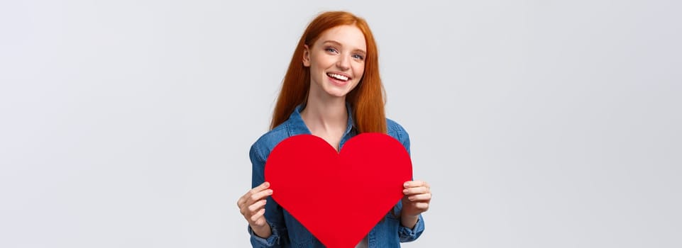 Valentines day, romance and teenagers concept. Lovely and cute redhead girlfriend asking wanna go prom together with big red heart, confessing love, smiling joyfully, white background.