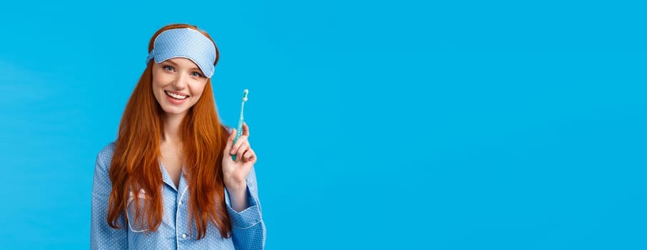 Health, beauty and lifestyle concept. Cheerful pretty european woman with blue eyes, toothy smile, holding toothbrush and smiling delighted, wearing sleep mask, nightwear, blue background.